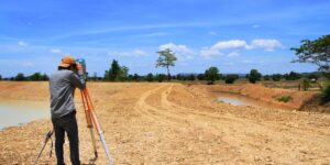 A man uses surveying equipment on a dirt path between two bodies of water under a blue sky.
