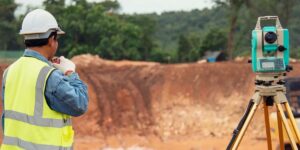 A surveyor in a hard hat and reflective vest uses a two-way radio next to a total station.