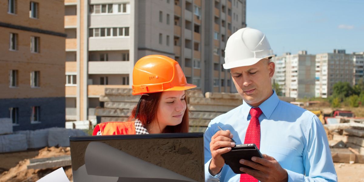 Two construction professionals, a man and a woman in hard hats, look at a tablet and blueprints outdoors