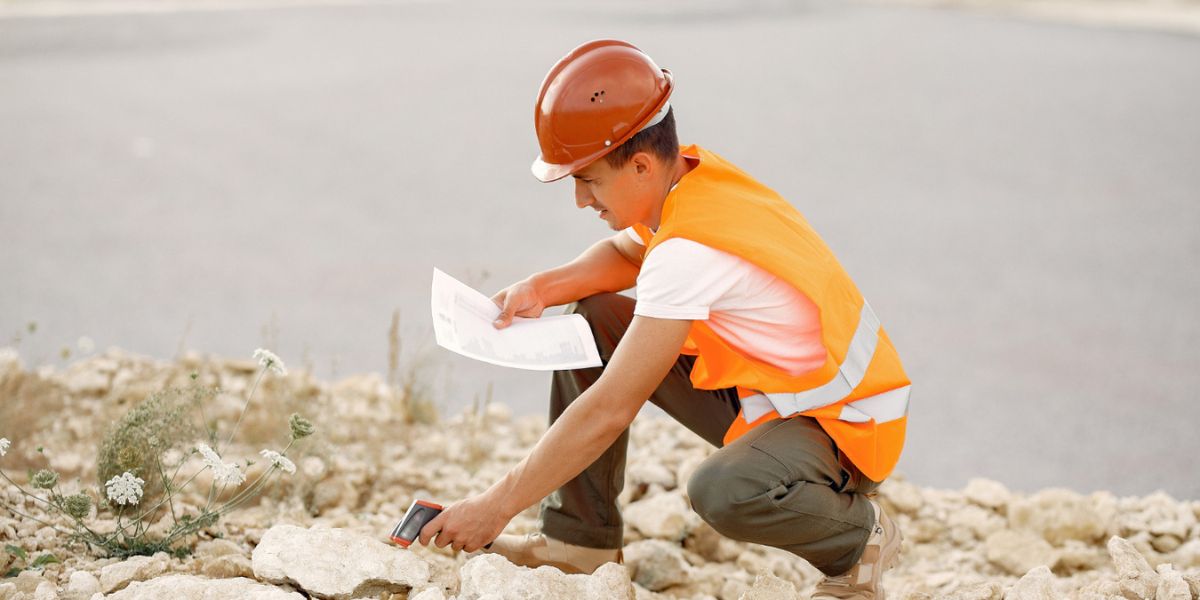 Young construction worker in a safety vest and hard hat crouches on rocky ground, examining documents